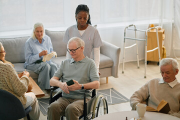 Group of elderly people sitting and engaging in activities while caregiver assisting one of them in...