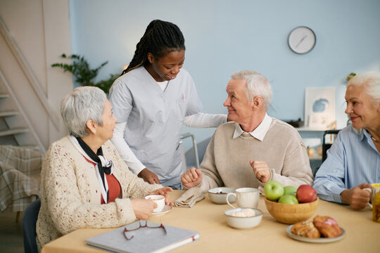 Caregiver engaging with elderly individuals while seated at dining table during meal, sharing stories and enjoying food together in cozy room with personal touches