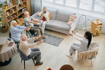 Group of elderly individuals participating in a seated exercise session inside comfortable living room with large windows and a walker nearby for support