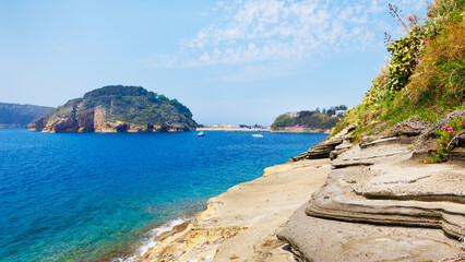 Rocky coastline of Procida island, Italy, with clear view of Punta della Palombara headland. Boats anchored in Mediterranean Sea and marina visible in background.
