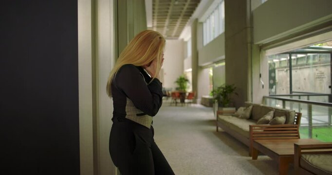 Woman covering part of face in emotional moment while leaning against wall in modern office hallway with couches and potted plants in soft blur