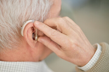 Close-up image of elderly person adjusting hearing aid behind ear, showing device clearly. Hand...