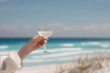 closeup of womans hand elegantly holding cocktail glass with beautiful beach in background