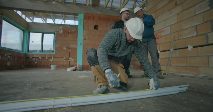 Construction worker measuring pipes with tape measure on floor, two colleagues reviewing blueprints in background, teamwork and planning in unfinished building interior