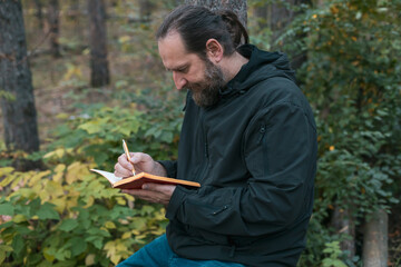 A man with a beard is sitting in a forest and writing in a notebook