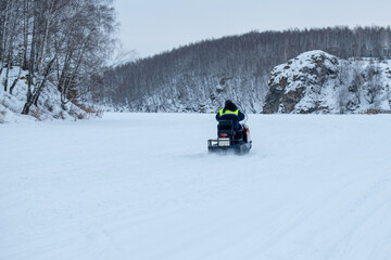 A man rides a snowmobile on a snowy road. The snowmobile is equipped with a snowplow, which is used to clear the road