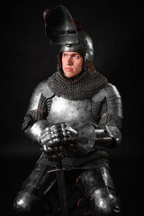 Naklejka premium Portrait of a knight in full plate armor holding a sword posing sitting on chair isolated on a dark background.