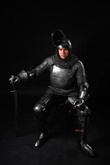 Portrait of a knight in full plate armor holding a sword posing sitting on chair isolated on a dark background.
