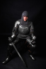 Portrait of a knight in full plate armor holding a sword posing sitting on chair isolated on a dark...