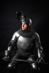 Portrait of a knight in full plate armor holding a sword posing sitting on chair isolated on a dark...