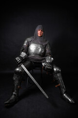 Portrait of a knight in full plate armor holding a sword posing sitting on chair isolated on a dark...