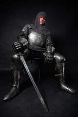Portrait of a knight in full plate armor holding a sword posing sitting on chair isolated on a dark...