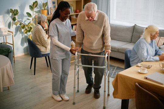 Nurse in uniform assisting senior man with walker inside cozy nursing home while another resident in background engages in activities