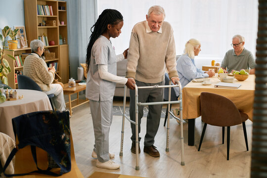Elderly man using walker receiving help from caregiver within senior care center as other residents engage in activities