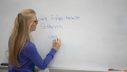 A middle-aged woman writes about complex inheritance patterns on a classroom whiteboard. She...