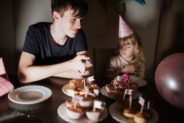 Siblings celebrate a birthday with cupcakes and party hats at home in the evening