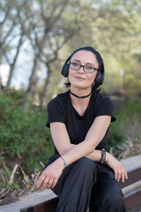 Young woman in black outfit relaxing on a bench, listening to music with headphones, eyes closed and smiling peacefully in a park on a sunny day