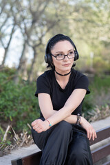 Stylish young woman in black clothes and headphones sitting on a bench in a park, enjoying music with a thoughtful expression on a sunny day