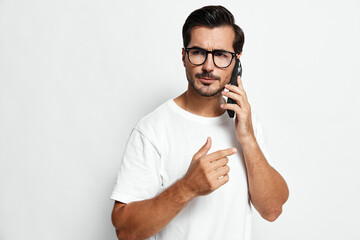Thoughtful man talking on mobile phone, wearing casual white t-shirt and glasses, looking at camera with serious expression isolated on plain light background.