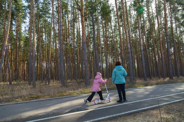 Little girl riding scooter while walking with her grandmother in the forest on a sunny day