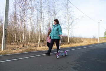 Little girl riding scooter while walking with her grandmother in the forest on a sunny day