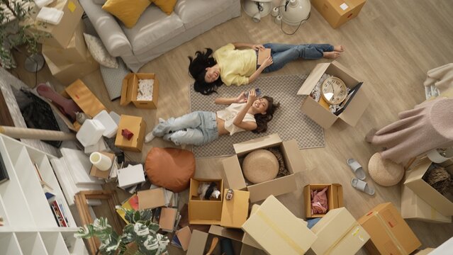 Lesbian lover looking at phone while laughing together and lying down at living room floor with cardboard box scattering. Happy couple checking at smart phone while spending time together. Myrmidon.