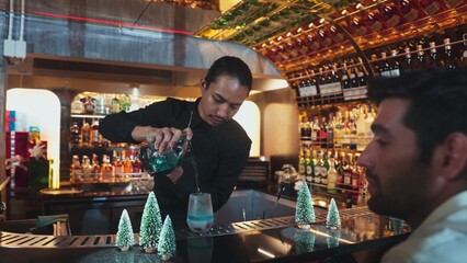 Bartender pouring mixed blue lagoon cocktail liqueur to cooling glass while guest talking friends waiting drink, decorated pine light christmas theme making special festive tasty in couple. Vinosity.