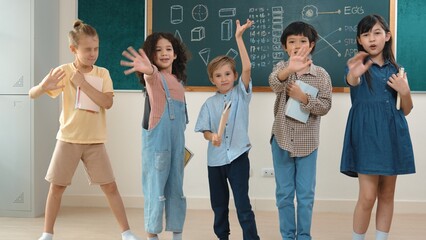 Fototapeta premium Multicultural elementary student in casual cloth looking at camera while waving hands at classroom. Group of diverse children hold notebook and smiling to camera at blackboard in classroom. Pedagogy.