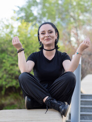 Smiling young woman in black streetwear sits cross-legged outdoors, hands raised in a playful meditation pose, surrounded by greenery on a sunny day