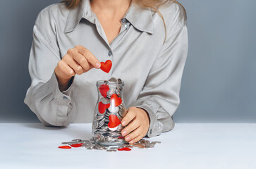 Donation jar with silver coins, into which hand of woman in gray shirt puts red heart. Sympathy...