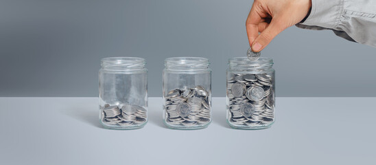 Three glass jars with coins as diagram. Female hand putting coin. Isolated on grey. Savings, finance, loans, pension concept.