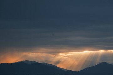Rays of Sunlight break through clouds above distant mountain peaks creating a serene and breathtaking landscape at sunset
