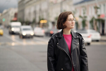 Young woman in a black leather jacket and pink sweatshirt walking down a city street. Confident street style look with casual fashion and urban atmosphere.