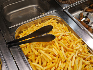 French fries, kept warm in a metal tray at a buffet, waiting to be served with black tongs, offer a delicious, crispy snack option