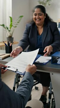 Smiling disabled plus-size indian businesswoman in wheelchair handing contract to businessman during office meeting
