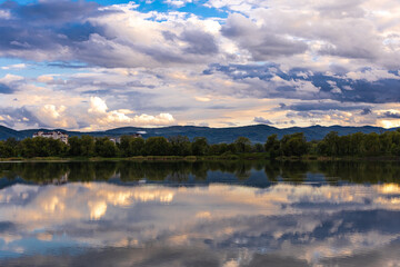 Serene lake view with a mountain silhouette and cloudy sky reflecting on water surface. Ideal for...
