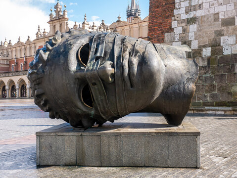 Igor Mitoraj Eros Bendato large head sculpture in Krakow Poland Main Market Square Rynek Glowny, Sukiennice historic architecture buildings in the background, outdoor bronze art installation, Europe
