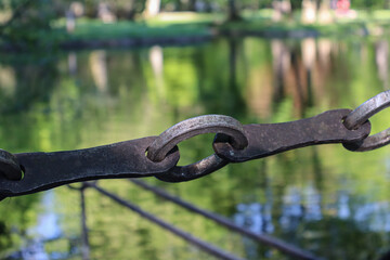 Close-up photo of a weathered iron chain link with green water and blurred reflections of trees in the background, rustic outdoor detail with shallow depth of field 