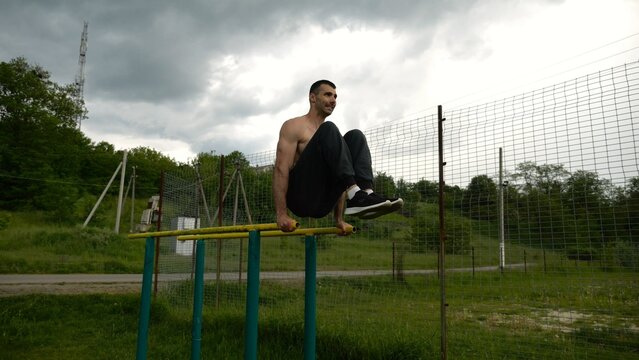Strong guy training abdominal muscles on parallel bars at sports ground. Young shirtless sportsman exercising at nature. Sporty athlete working out at nature. Concept of active sportive lifestyle