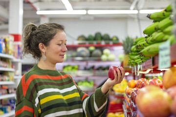Smiling female customer is choosing fresh produce, holding red apple in hand, buying groceries in...