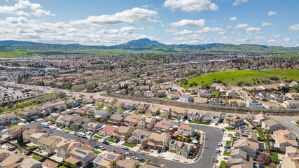 Aerial view of a suburban neighborhood and shopping center in Antioch, California, on a clear spring day with scenic hills, green fields, and residential homes with solar panels.