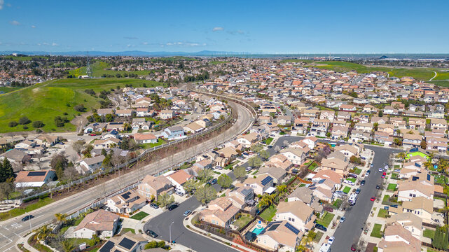Aerial view of a suburban neighborhood and shopping center in Antioch, California, on a clear spring day with scenic hills, green fields, and residential homes with solar panels.
