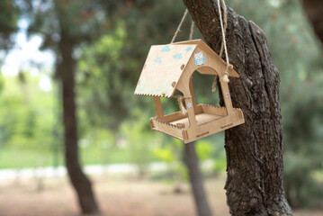 A wooden birdhouse hangs from a tree branch in a park during springtime. The surrounding area features green foliage and soft sunlight filtering through the leaves.