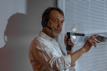 Detective on assignment. A man in a headset stands at a window with blinds, conducting covert...