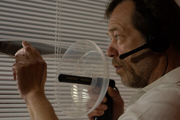 Spying in action. A man uses professional eavesdropping equipment, peering through the blinds.