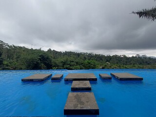 Stone stepping stones in a tranquil blue pool against a lush green forest and cloudy sky creating a serene landscape