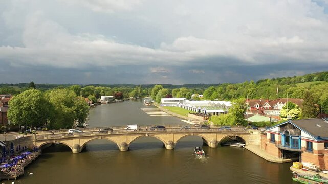 Aerial drone footage flying towards and over the historic Henley Bridge, revealing the River Thames, riverside town buildings, and event marquees set up along the bank under a cloudy sky.