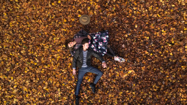Aerial view of an attractive young woman and man, while laying on the forest floor or autumn leaves carpet. Elevated photo of romantic couple enjoying love and fall forest.