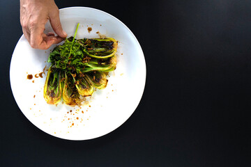 Hand adds finishing touches to a plate of bok choy with herbs in a stylish kitchen setting.