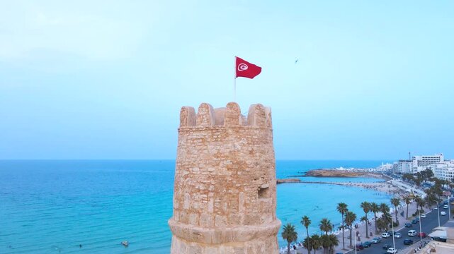 A picturesque scene featuring a historic stone tower flying a flag, set against a vibrant Mediterranean background with a clear blue sky and calm seas.